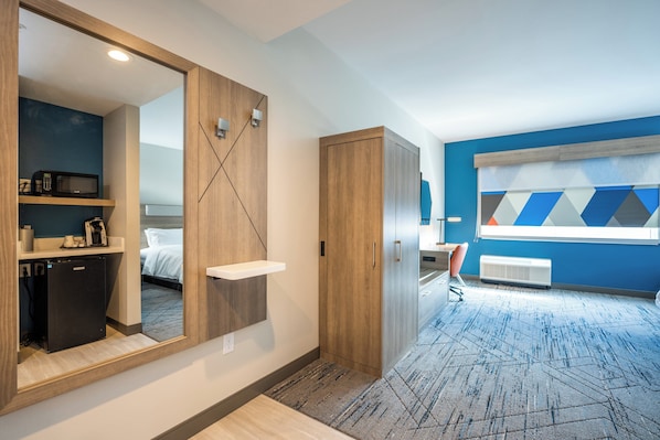 A modern hotel room with a large mirror, wooden wardrobe, a small kitchenette, and a blue accent wall.