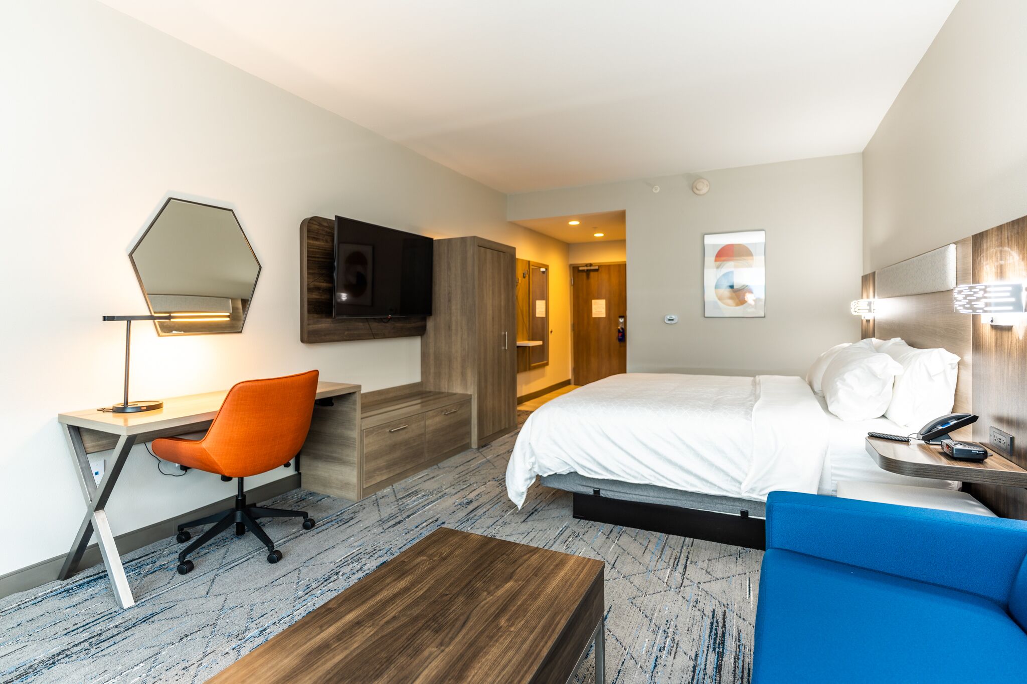 Une chambre d’hôtel avec un lit, un bureau et une chaise, un canapé bleu, une table basse en bois, une télévision et un miroir au mur.