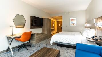 A hotel room with a bed, a desk with a chair, a blue sofa, a wooden coffee table, a TV, and a mirror on the wall.