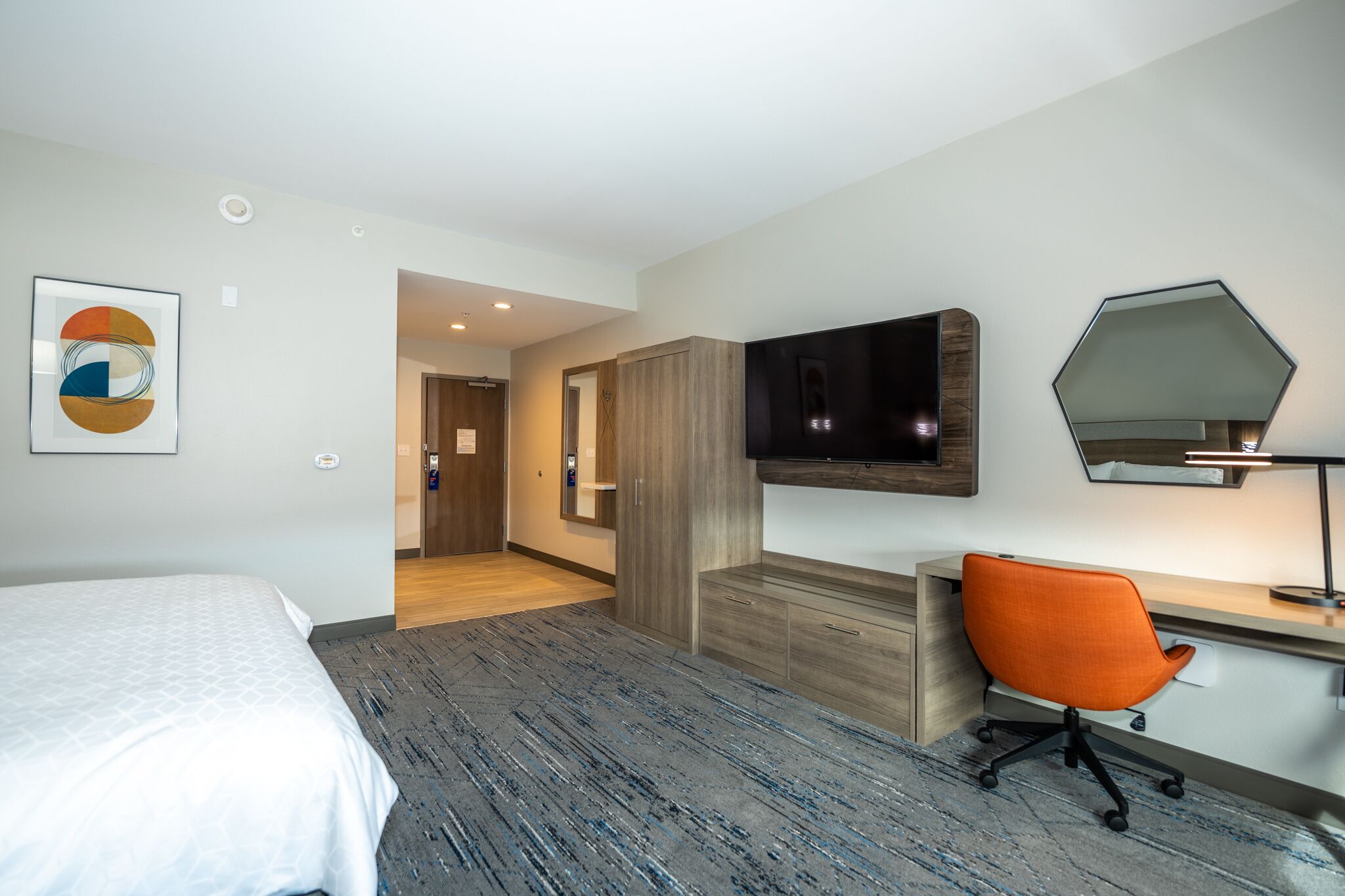 Habitación de hotel con una cama, un escritorio con una silla, un televisor y un espejo.