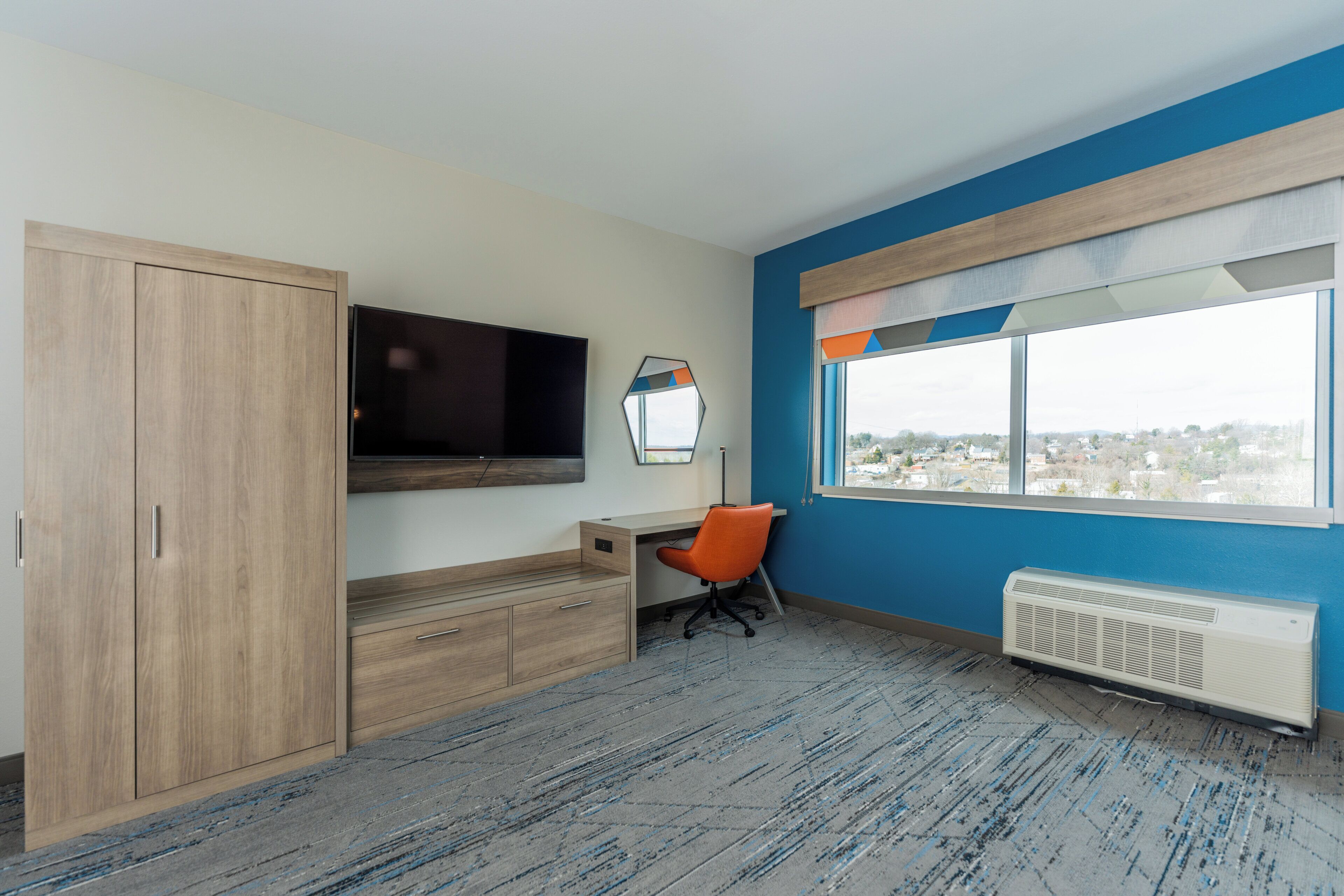 A room with a large wardrobe, a flat-screen TV mounted on the wall, a desk with an orange office chair, and a window with a view of buildings.