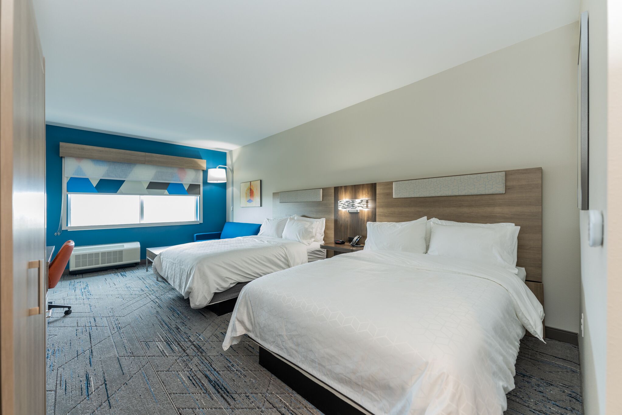 Habitación de hotel con dos camas, un escritorio y una silla. Se ve una ventana con cortinas azules y una obra de arte en la pared.