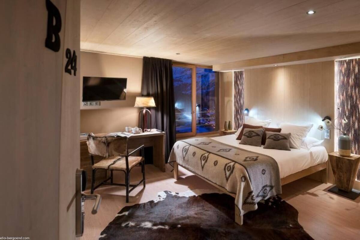 A modern hotel room with a large bed, a desk with a chair, a television, and a view of the night sky through the window.