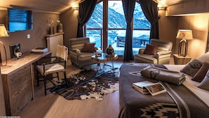 Een knus ingerichte kamer met een houten plafond, een groot raam met gordijnen, een bed, een bureau met stoel en een klein tafeltje met lamp.