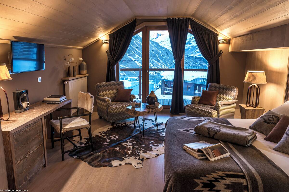A cozy room with a wooden ceiling, a large window with curtains, a bed, a desk with a chair, and a small table with a lamp.