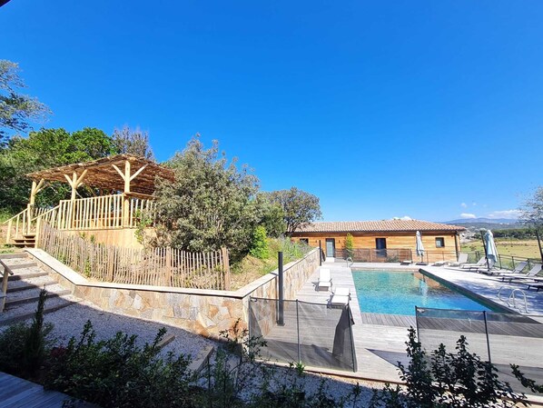 Ein Poolbereich mit Holzterrasse, einer Pergola und einem Gebäude mit Ziegeldach.