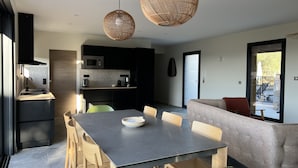 A modern kitchen with dark cabinetry, a microwave, and a coffee maker. There is a dining table with chairs and a woven pendant light. A sofa is visible in the background, and a door leads to an outdoor area.
