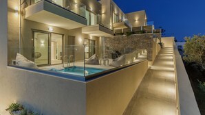 Une maison moderne à plusieurs étages, dotée d’une piscine intérieure à panneaux de verre, d’un espace de détente extérieur et d’un mur de soutènement en pierre.