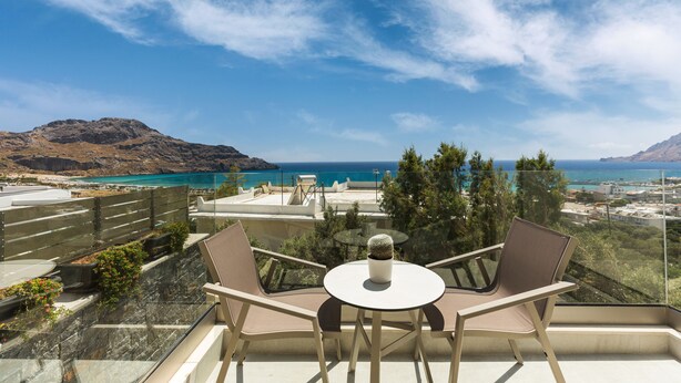 Un balcon avec deux chaises et une table, donnant sur un paysage côtier avec des montagnes et un ciel bleu dégagé.