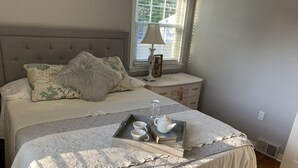 A neatly made bed with a quilted bedspread, a bedside table with a lamp, a tray with a teapot and cups, and a window with blinds. - Buffalo Ny Carols Cape (Bowmansville)