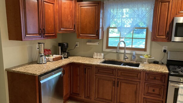 A kitchen with wooden cabinets, a stainless steel dishwasher, a sink, a stove, and a window with a view of greenery. - Buffalo Ny Carols Cape (Bowmansville)