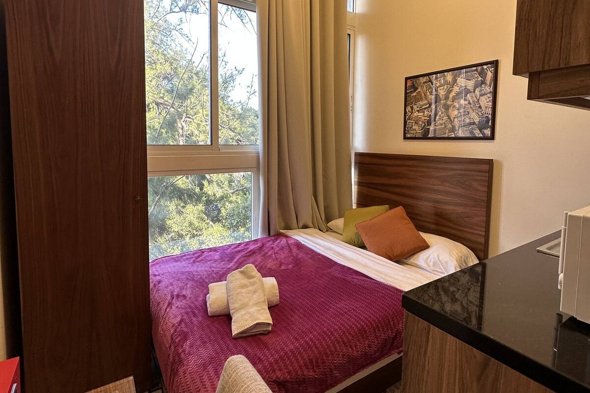 A hotel room with a bed, a desk, a chair, a window with curtains, and a picture on the wall.