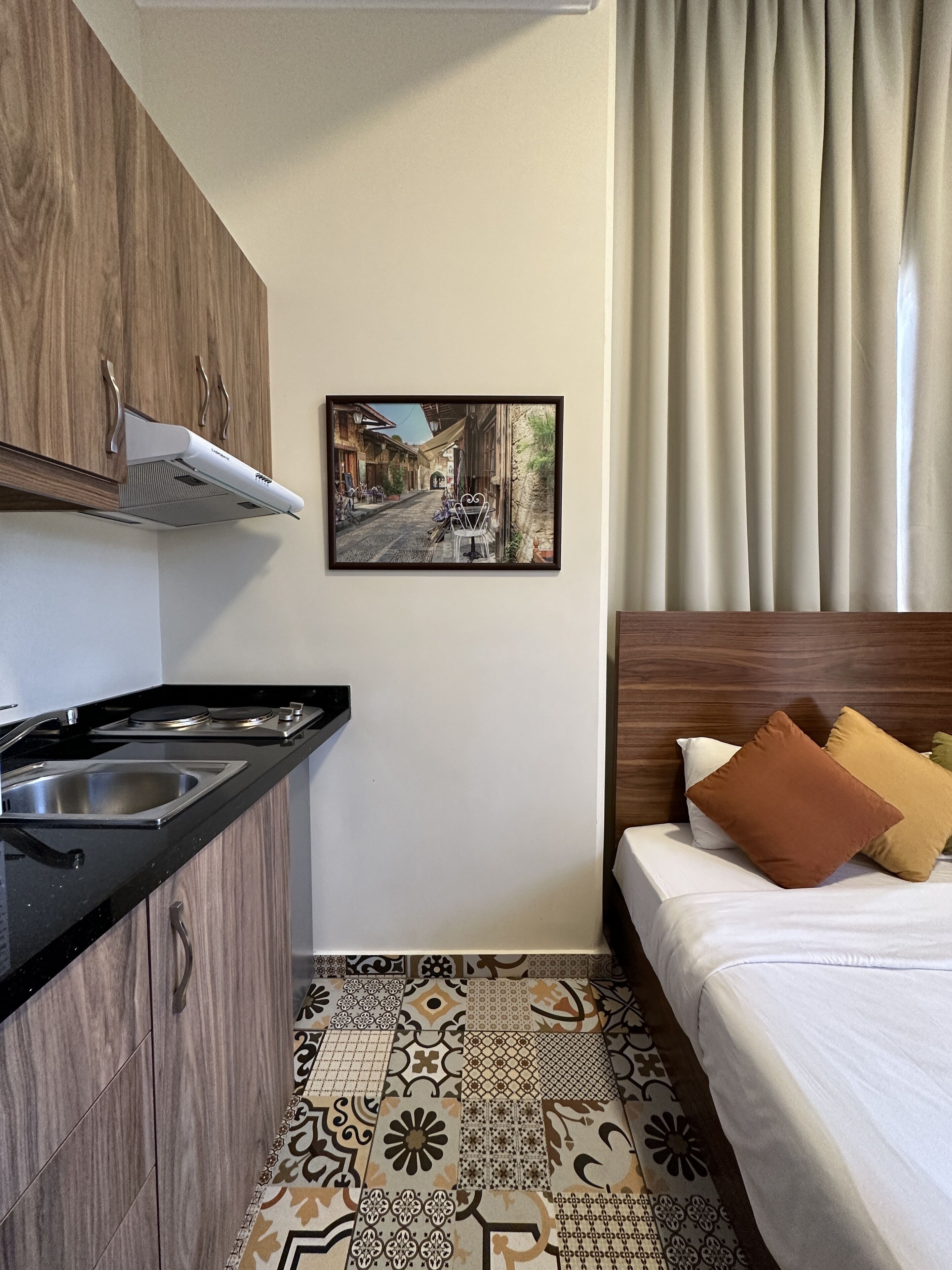 Ein kleines Hotelzimmer mit Küchenzeile, einem Bett mit Kissen, einem gemusterten Boden und einem gerahmten Bild an der Wand.