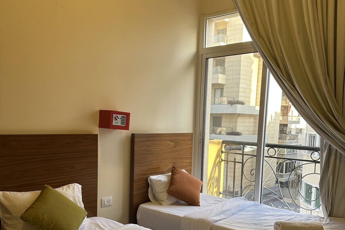 A hotel room with two beds, a wooden headboard, a window with curtains, and a view of buildings outside.