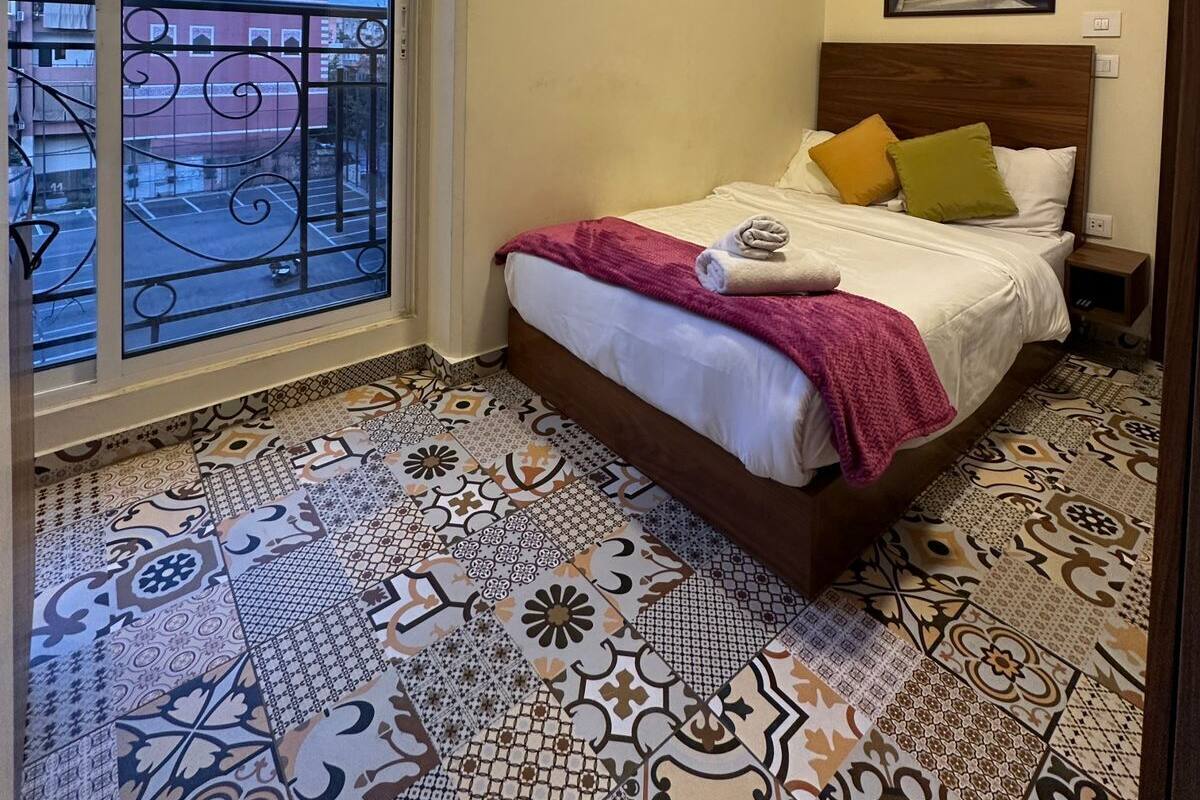 A bedroom with a large window, a bed with pillows, and a patterned floor.