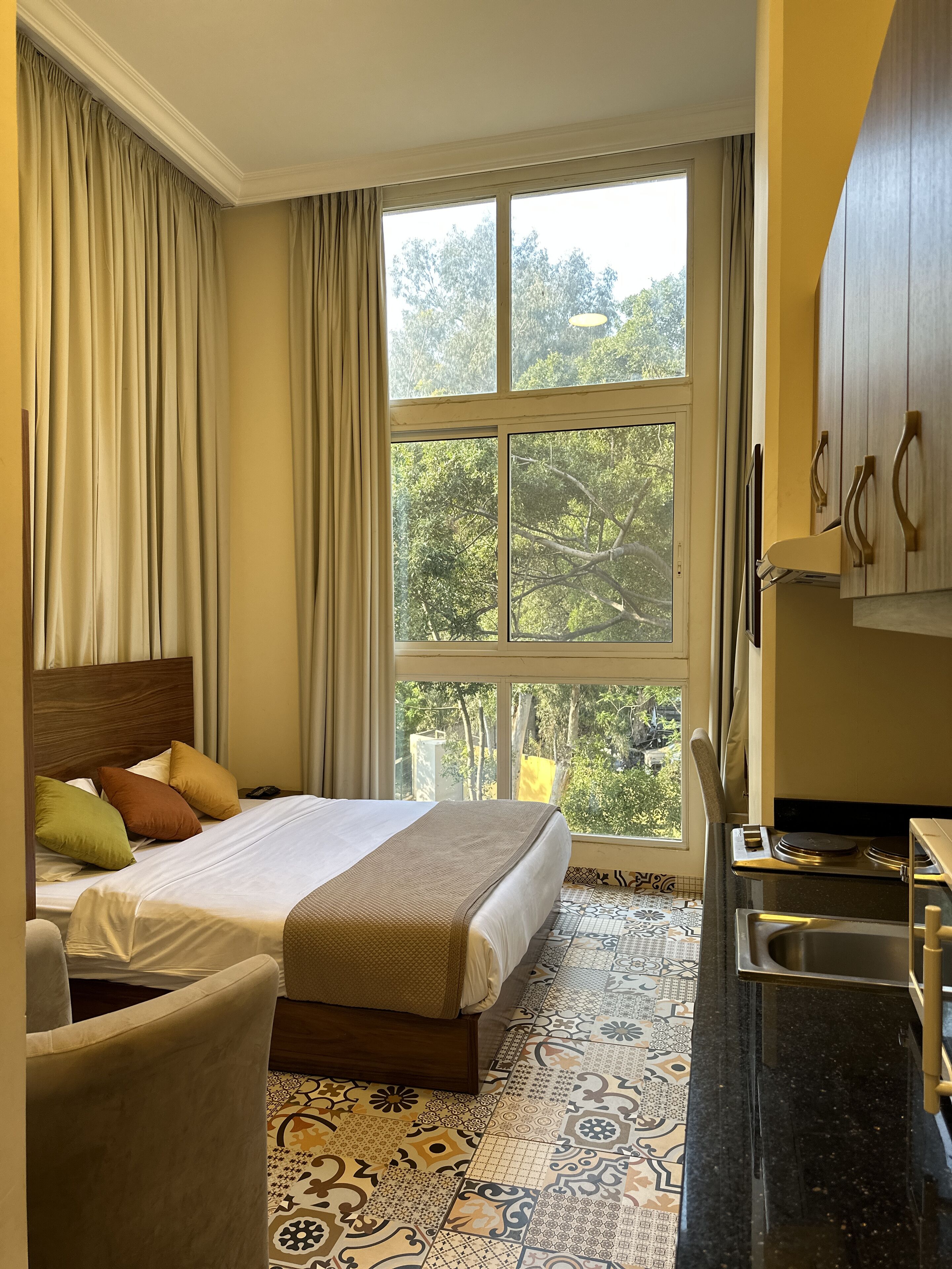 A hotel room with a bed, a chair, a kitchen area, and a large window with a view of trees.