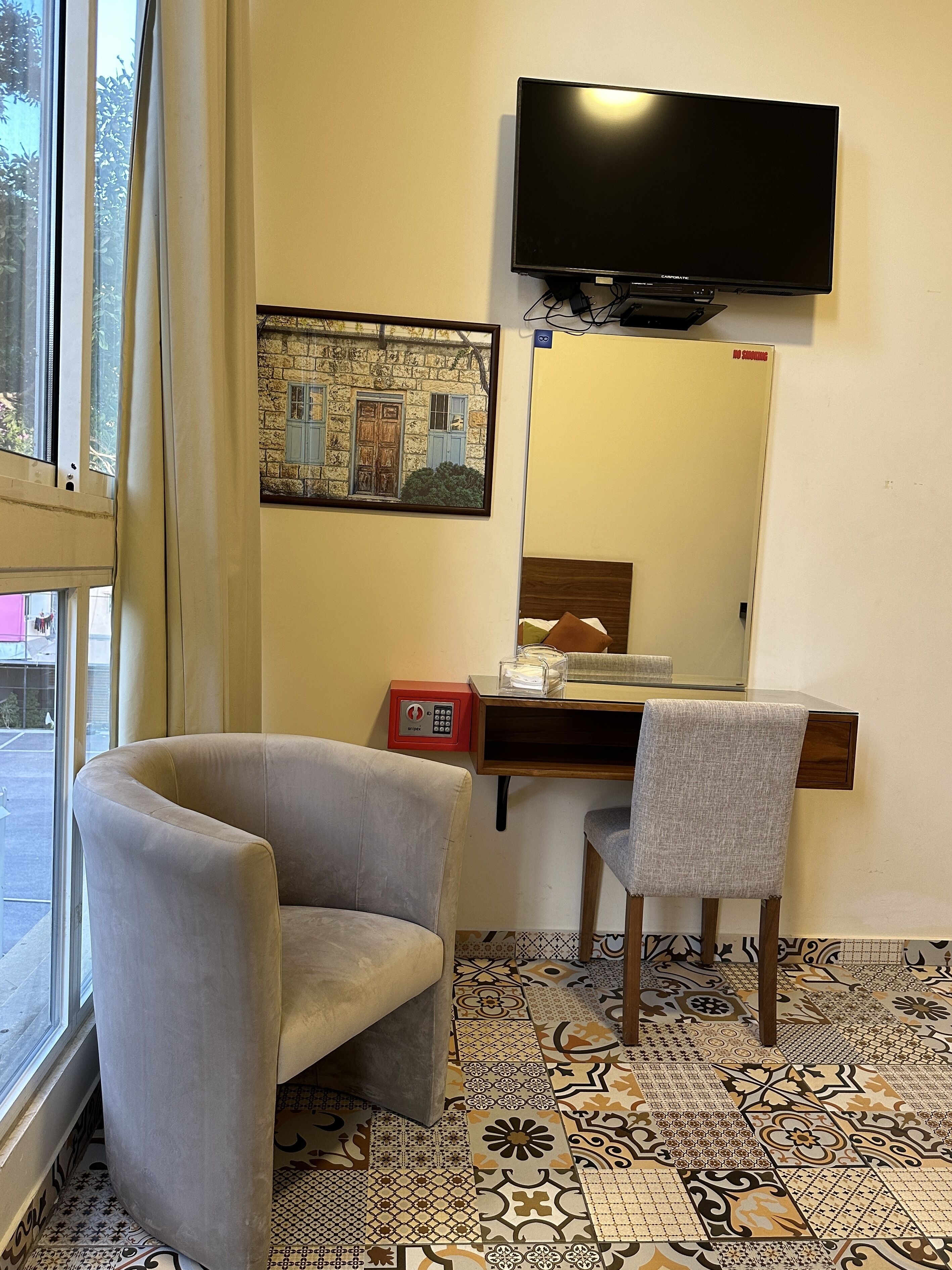 Une chambre d’hôtel dotée d’une grande fenêtre, d’un téléviseur à écran plat fixé au mur, d’un tableau encadré représentant un bâtiment, d’un bureau avec une chaise et d’un sol à motifs.
