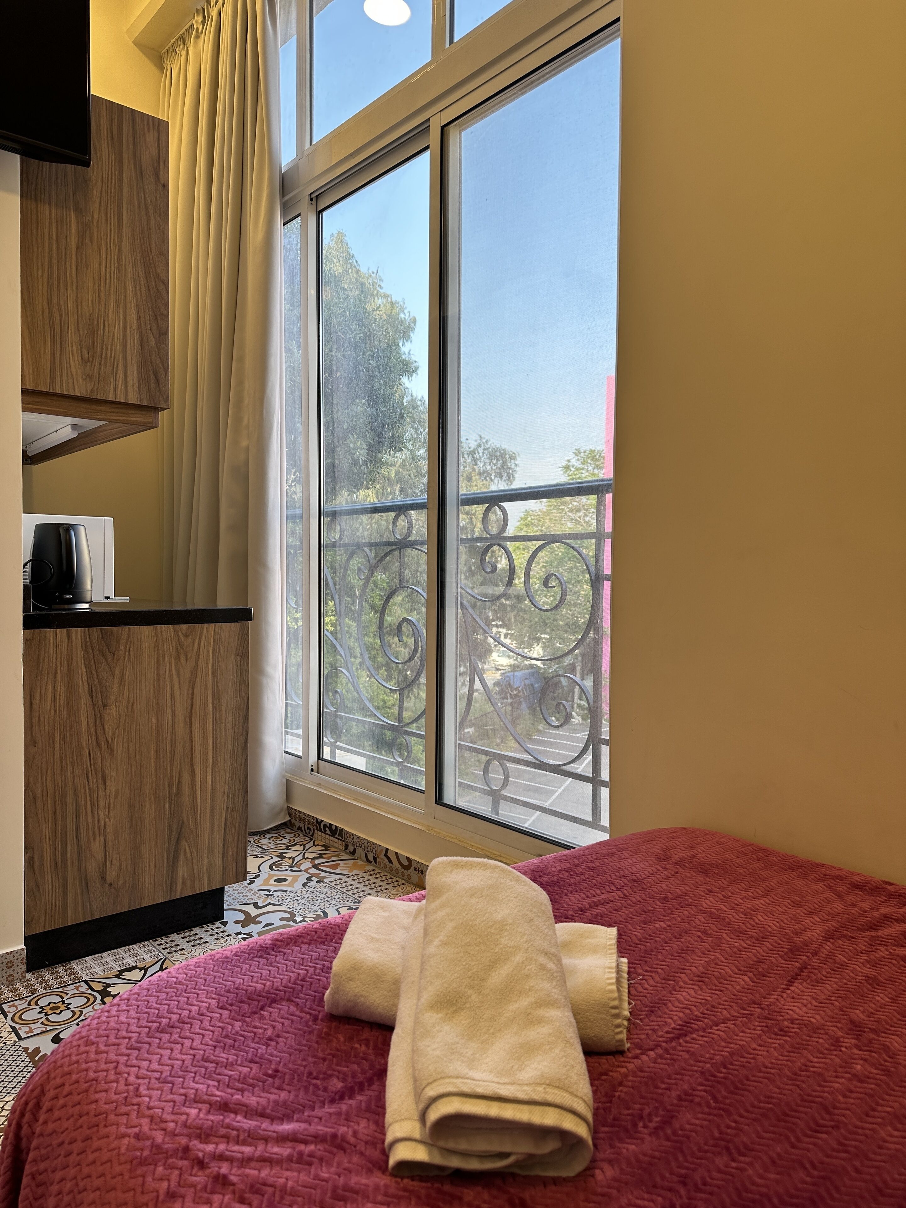 A bedroom with a large window, a bed with a red blanket, and a wooden cabinet.
