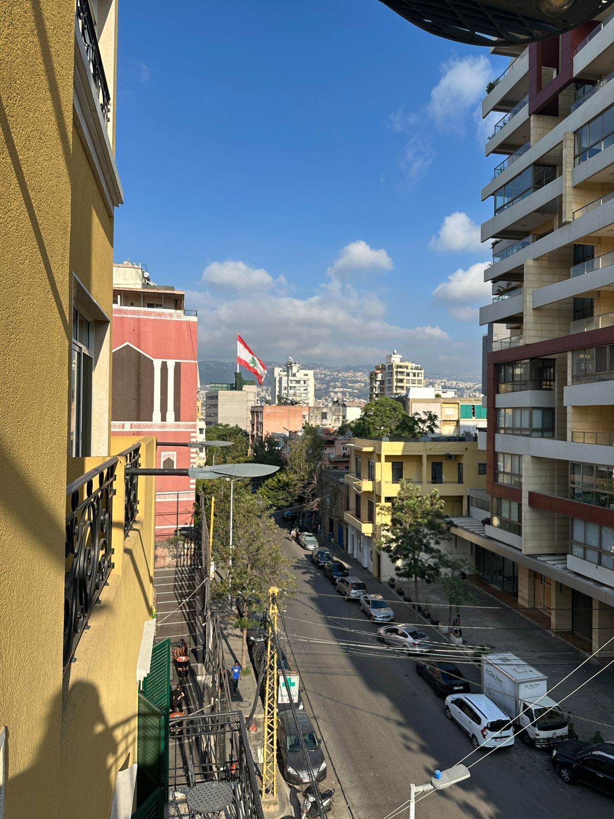 Un edificio de varios pisos con balcones y vistas a una calle con coches estacionados y una bandera.