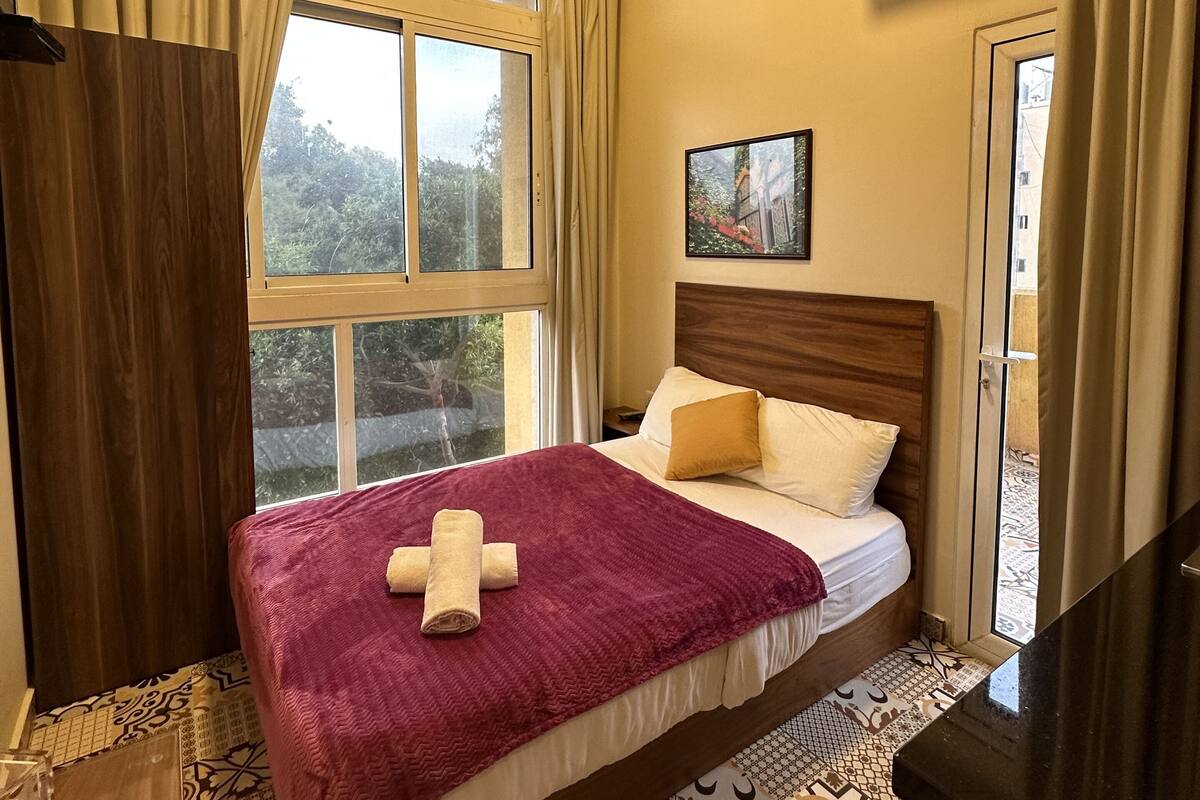 A hotel room with a large bed, a wooden headboard, a window with curtains, a small bedside table, and a picture on the wall.