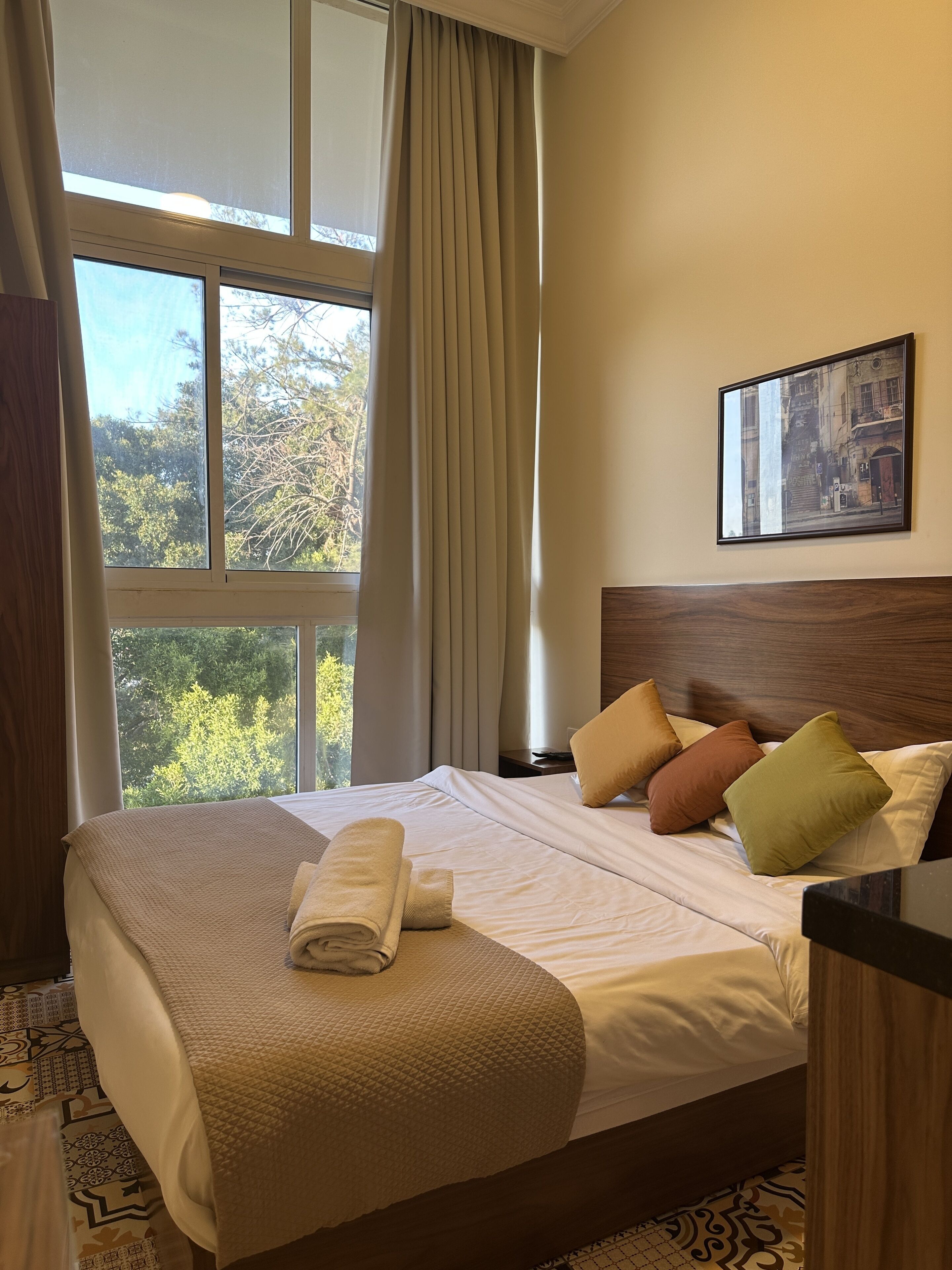 Ein Hotelzimmer mit einem großen Bett, einem Fenster mit Vorhängen, einem gerahmten Bild an der Wand und einem ordentlich bezogenen Bett mit einem aufgerollten Handtuch.
