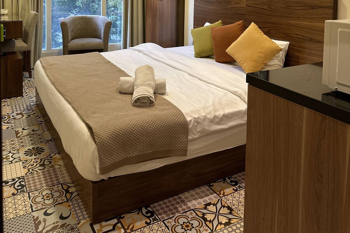 A hotel room with a large bed, a wooden wardrobe, a desk, a chair, and a large window with curtains.