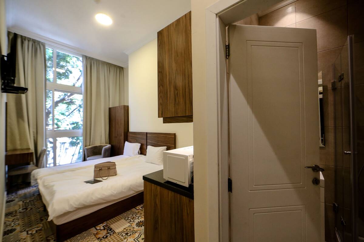 A hotel room with a bed, a bedside table, a wardrobe, a window with curtains, and a door.