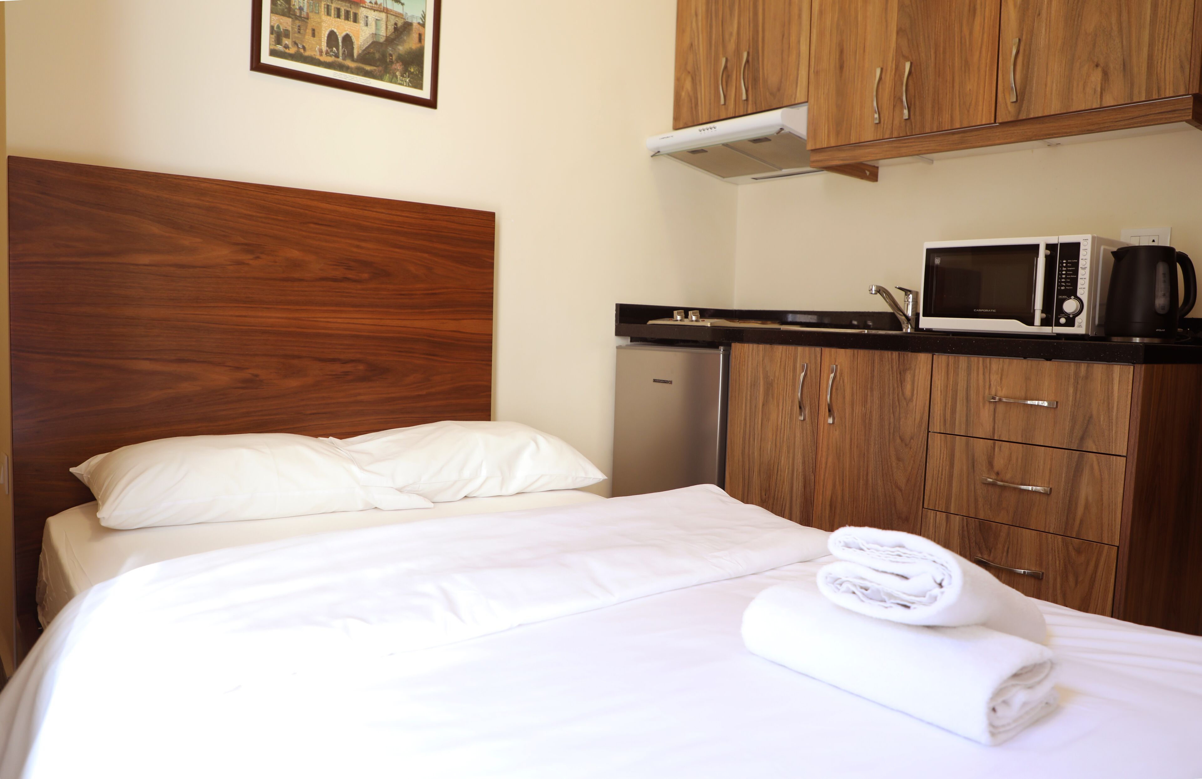 A hotel room with a bed, a wooden headboard, a microwave, a kettle, and a small refrigerator.