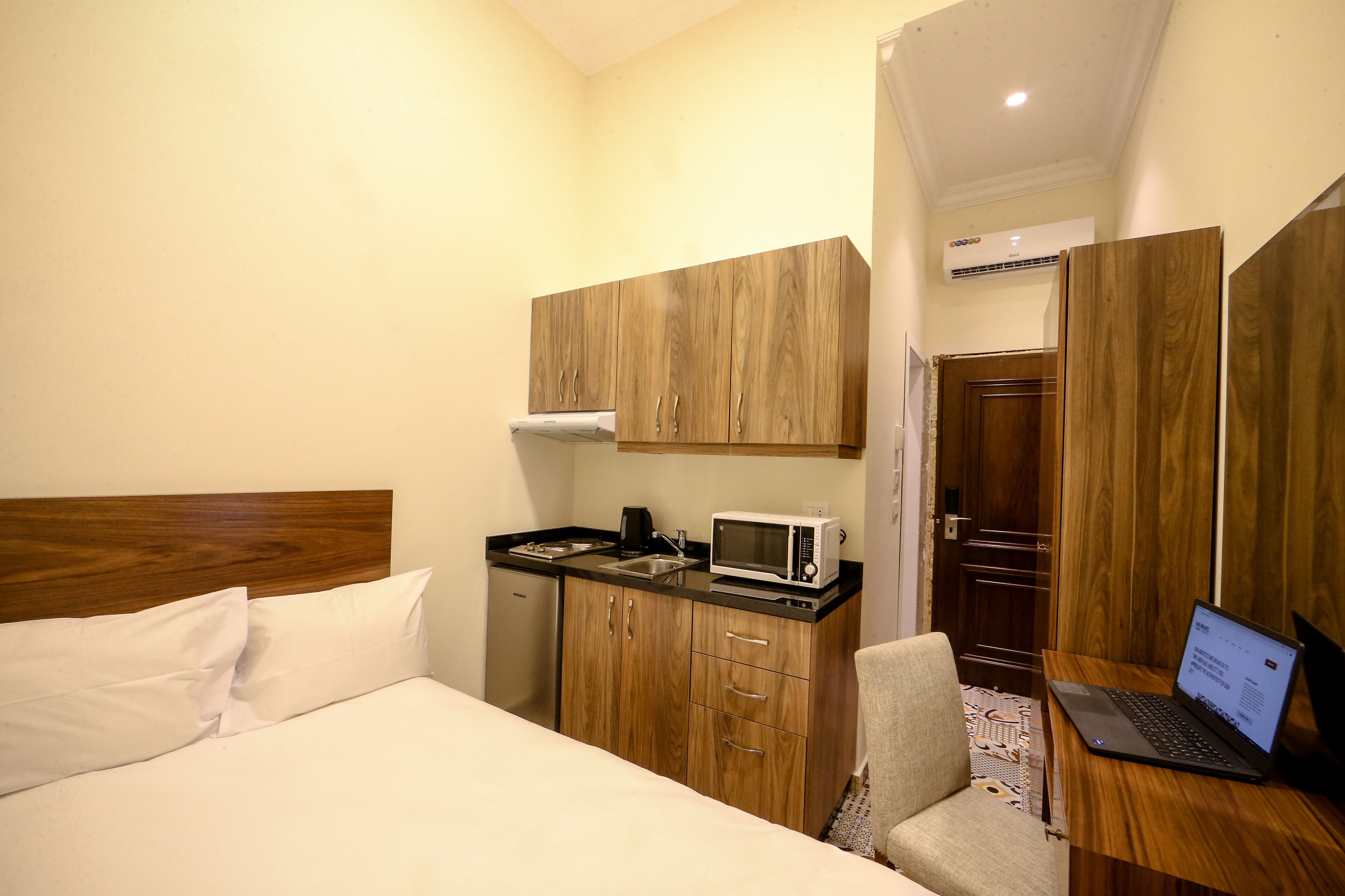 Une chambre d’hôtel compacte comprenant un lit, une kitchenette, un bureau avec un ordinateur portable et une porte donnant sur une autre pièce.