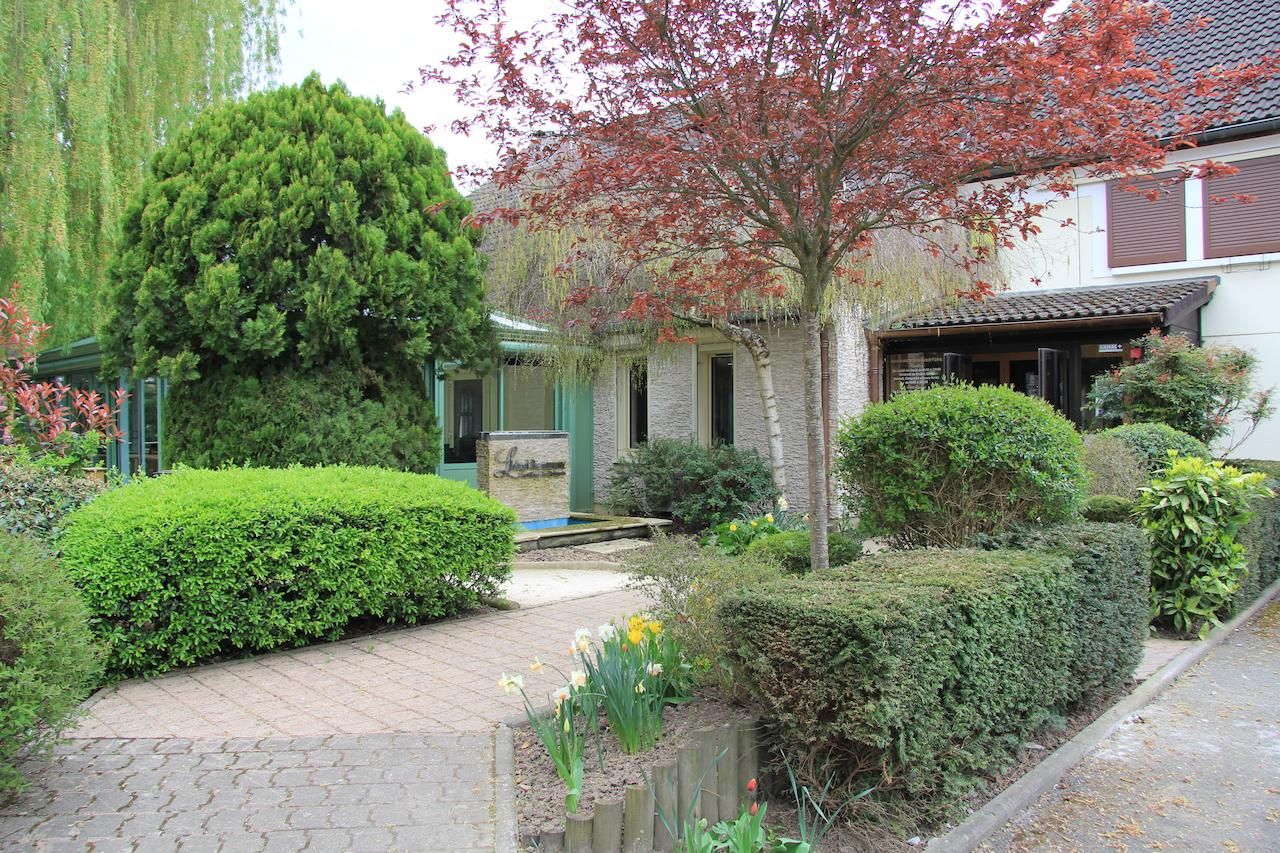 Une maison traditionnelle de style japonais, dotée d’un jardin bien entretenu comprenant un étang, divers arbustes et un grand arbre aux feuilles rouges.