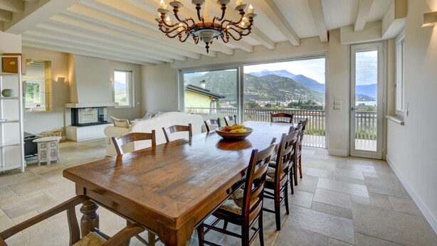 Un comedor amplio con una gran mesa de madera y sillas, una araña y vistas a montañas a través de amplios ventanales.