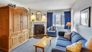 Ein Wohnzimmer mit Kamin, einem blauen Sofa, einem Holzkabinett und einem großen Fenster mit Vorhängen.