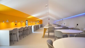 Una sala de conferencias moderna con mesas y sillas blancas, una mesa larga con sillas, y una pared de acento amarilla.