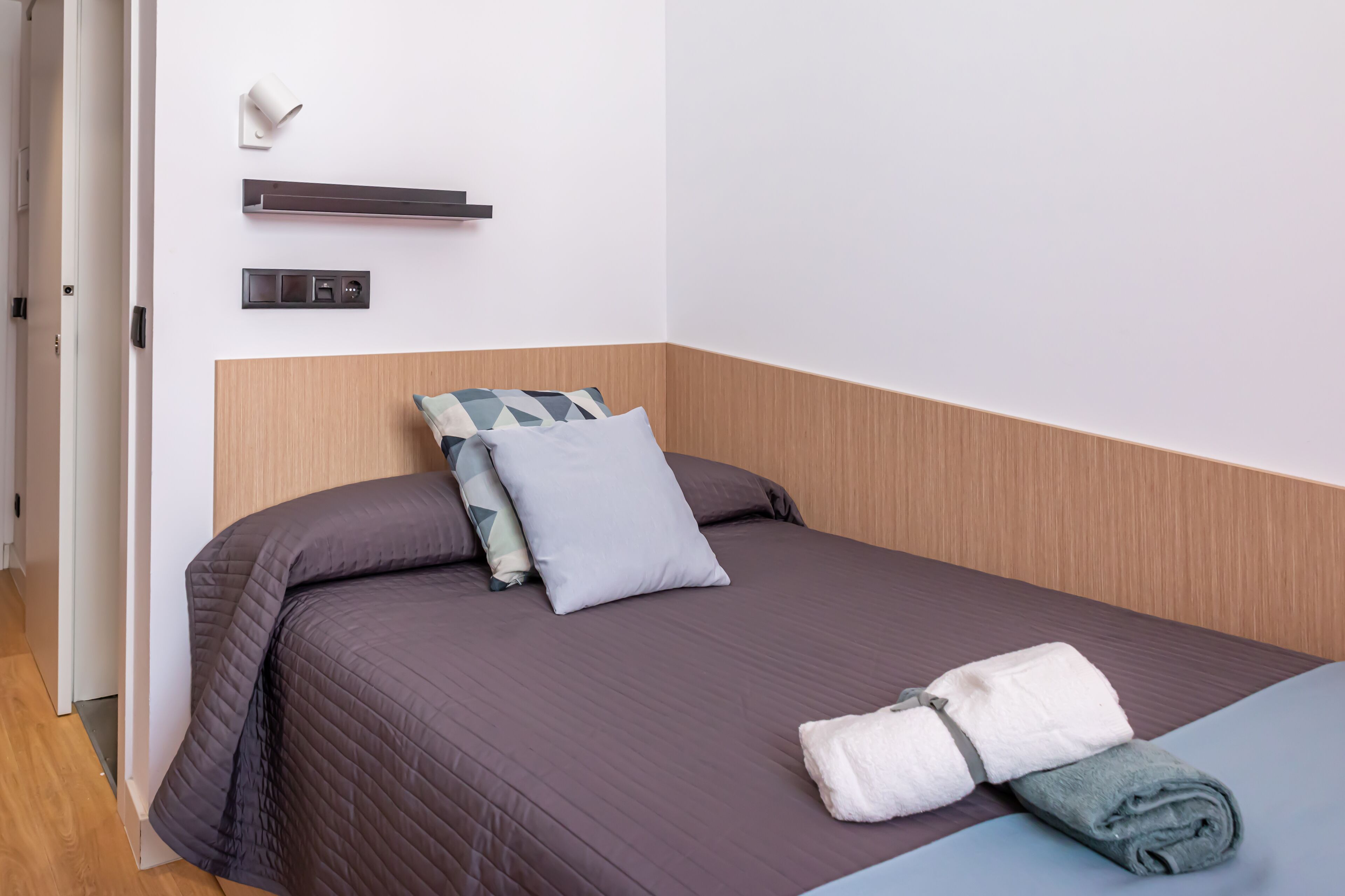 A neatly made bed with a dark quilt, two pillows, and a folded towel on a blue mattress.