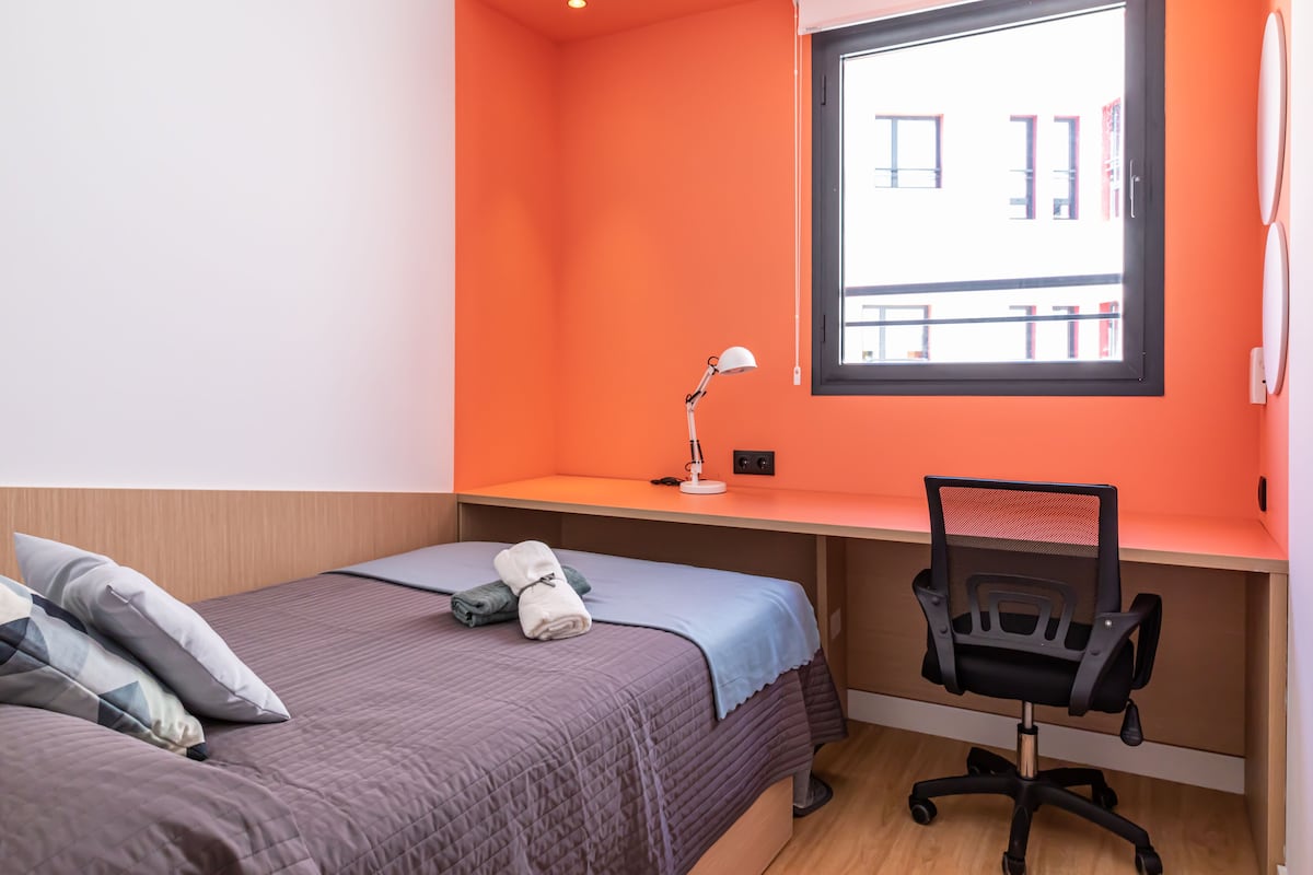 Um quarto com uma cama, uma escrivaninha, uma cadeira e uma janela.
