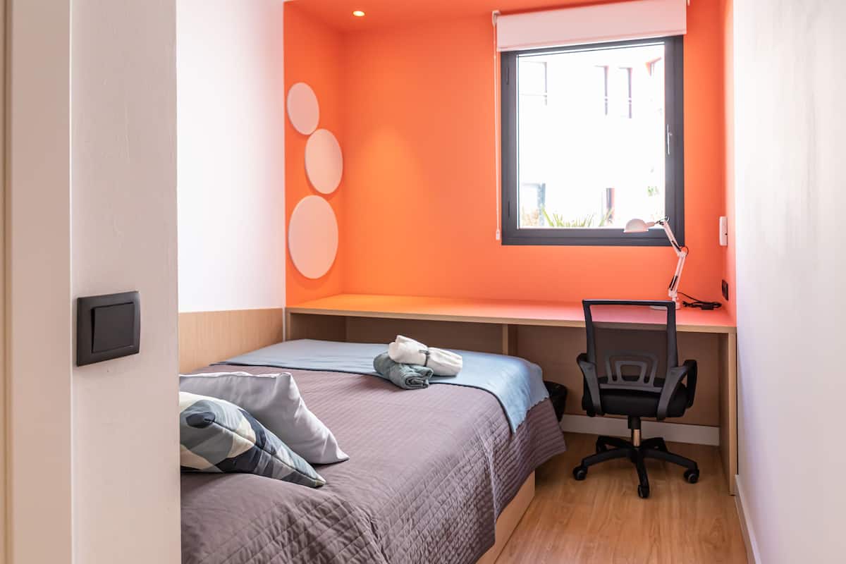 Quarto compacto com duas camas, uma escrivaninha e uma janela.