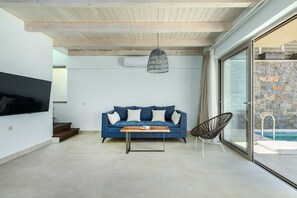 A modern living room with a blue sofa, a wooden coffee table, a hanging light fixture, and a glass door leading to a pool area.