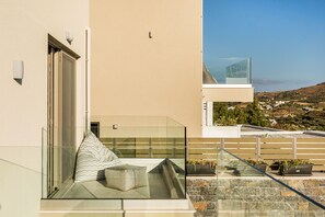 A modern balcony with a glass railing, a cushioned seating area, and potted plants.