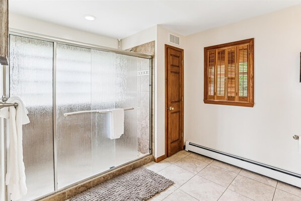 A bathroom with a glass shower enclosure, a wooden door, and a window with wooden shutters.