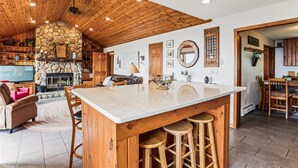 Een ruime keuken met een groot eiland, houten plafond, stenen open haard en een eetgedeelte met houten stoelen en krukken.