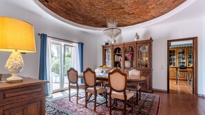 A dining area with a wooden table, chairs, and a chandelier. There is a large window with blue curtains and a view of greenery outside. - Villa with 7 bedrooms and private pool in Alporchinhos, walking distance to beach (Porches)