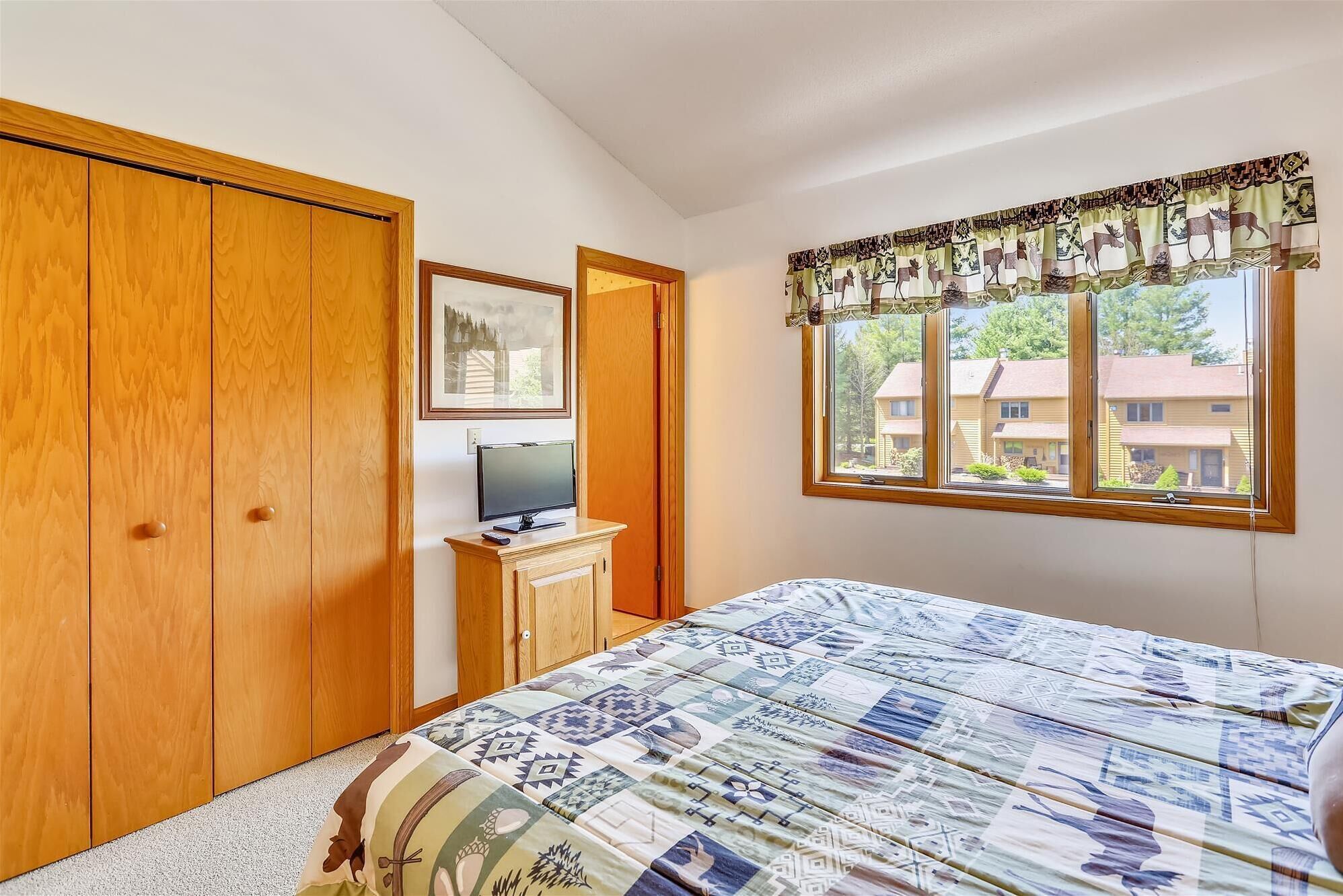 A bedroom with a bed, a TV on a stand, wooden wardrobe, and a window with a view of houses.