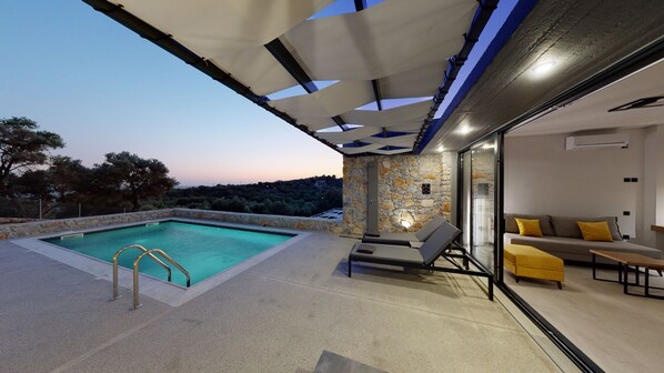 A modern outdoor pool area with a stone wall, lounge chairs, and a glass door leading to a living area with a sofa and yellow cushions. - DAELIA VILLAS II of 3 Luxury Villas, private pool, in an enclosed olive grove (Plomari Lesvos)