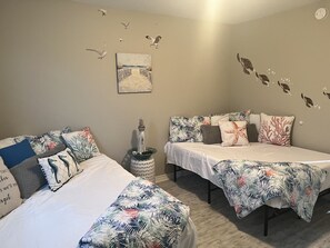 Ein Schlafzimmer mit zwei Betten, jedes mit einer Bettdecke mit tropischem Blattmuster, einem kleinen dekorativen Leuchtturm und Wandbildern mit Möwen und einer Strandansicht.