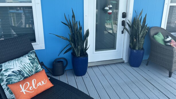 Une façade bleue avec une porte blanche, des plantes en pot, un fauteuil en osier et un espace de détente agrémenté d’un coussin sur lequel est inscrit le mot «Détente».