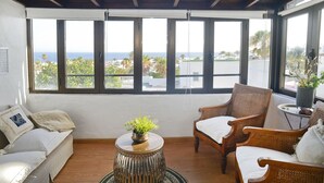 Ein Zimmer mit Holzboden, großen Fenstern mit schwarzen Rahmen, einem weißen Sofa, zwei Korbstühlen, einem kleinen runden Tisch mit einer Pflanze und Blick auf Palmen und das Meer.