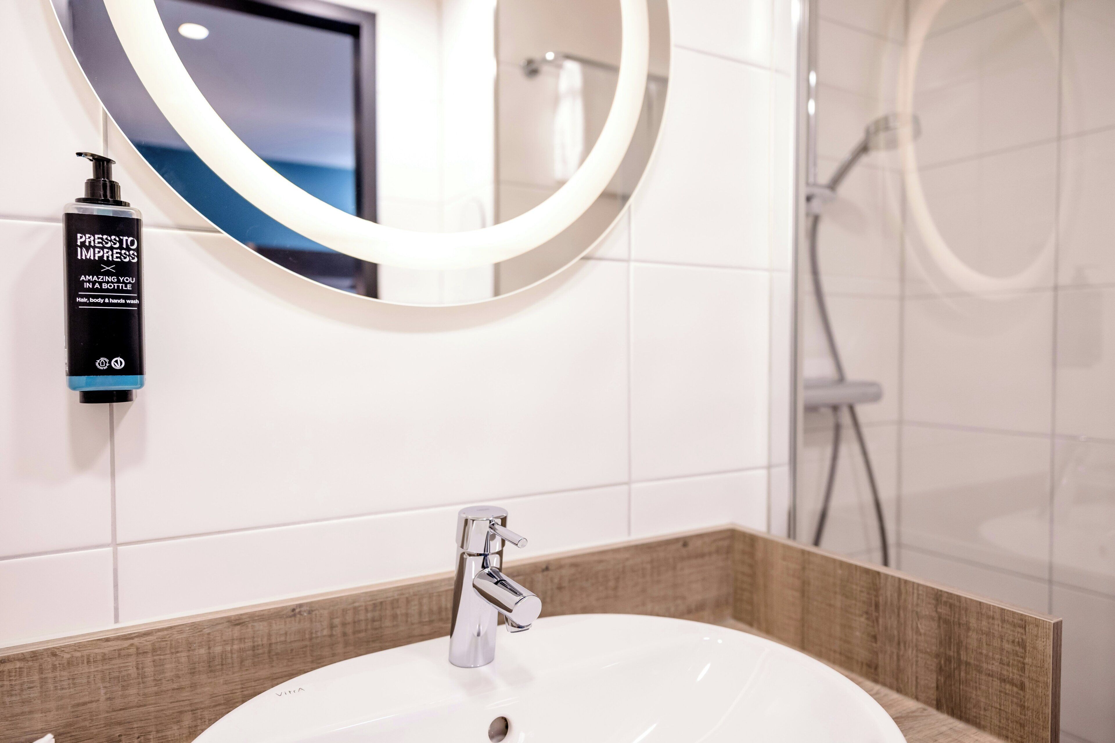 A bathroom with a white sink, a round mirror, a handheld soap dispenser, and a shower area.