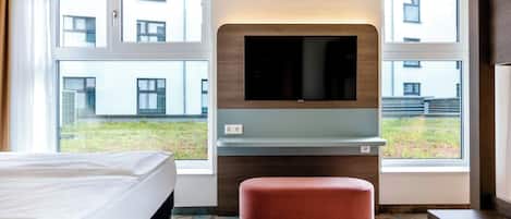Une chambre d’hôtel avec un lit, un téléviseur fixé au mur, un petit coin salon et de grandes fenêtres.