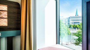 A room with a large window, a bench, a wall-mounted light, and a view of a building with a spire. - Hotel Kiel-Schwedenkai (Kiel)