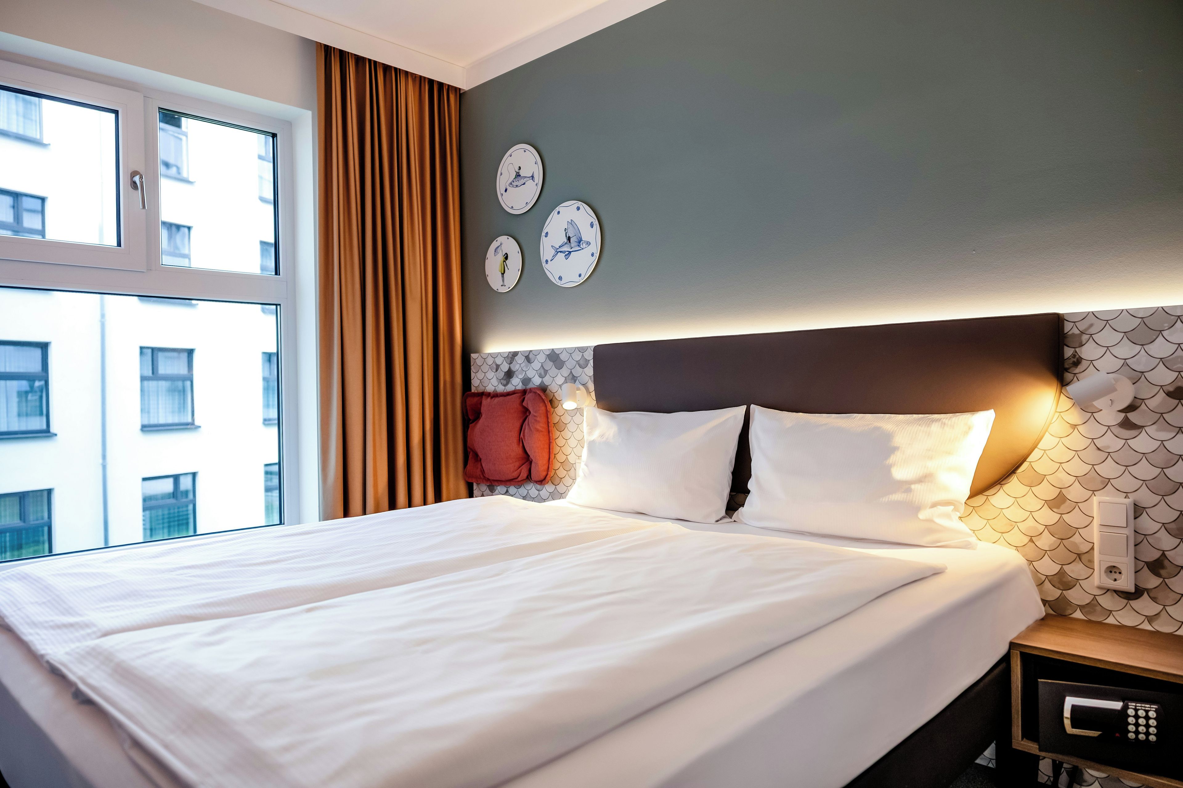 A hotel room with a bed, curtains, wall clocks, and a nightstand with a remote control.